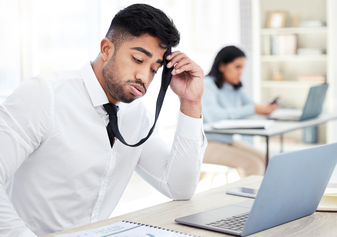 Stressed salesperson sitting at desk using his tie to wipe his forehead